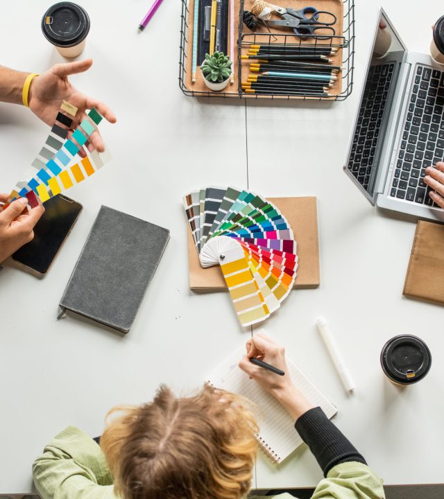 Overview of three young creative designers sitting by desk and choosing colors for new project from palette while one of them making notes